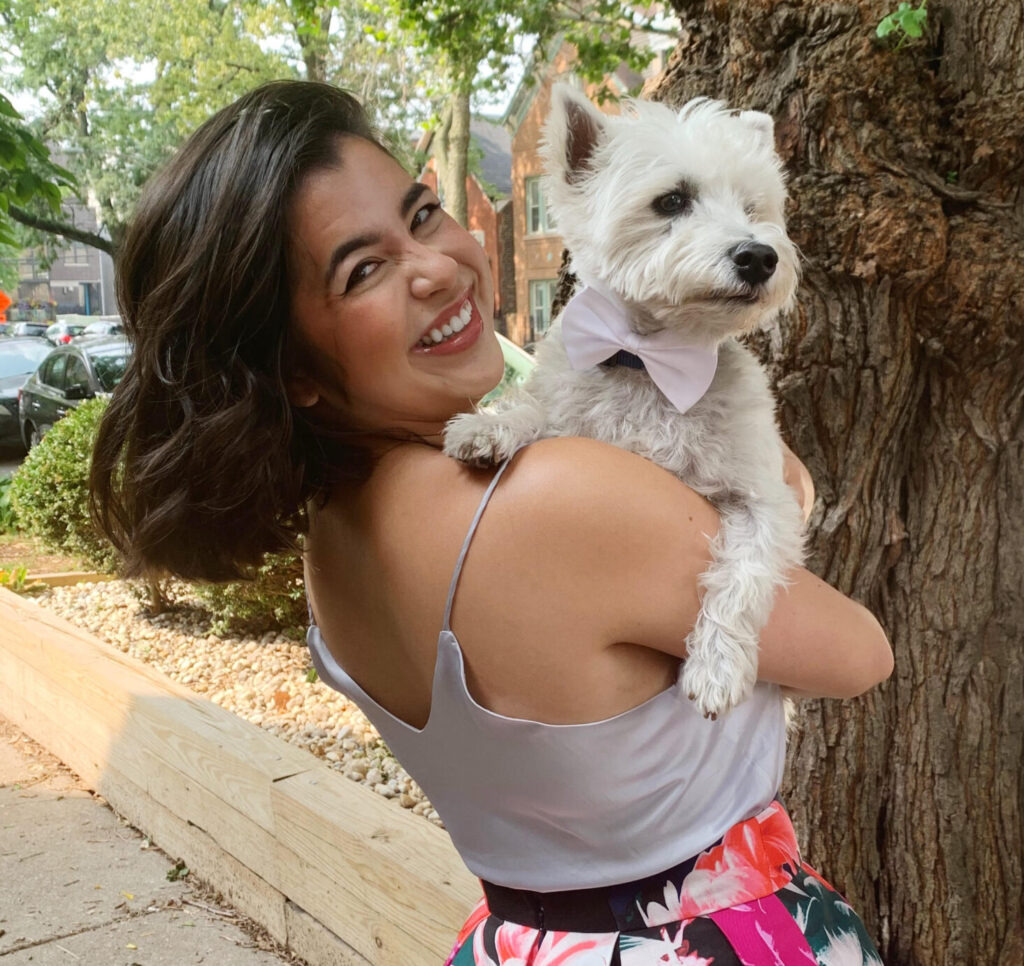 Erin is a young woman with dark brown hair. She is holding a small white dog and smiling at the camera.