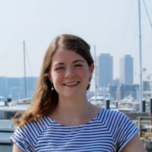 Libby is a young woman with brown hair standing in front of a city skyline. She is smiling at the camera.